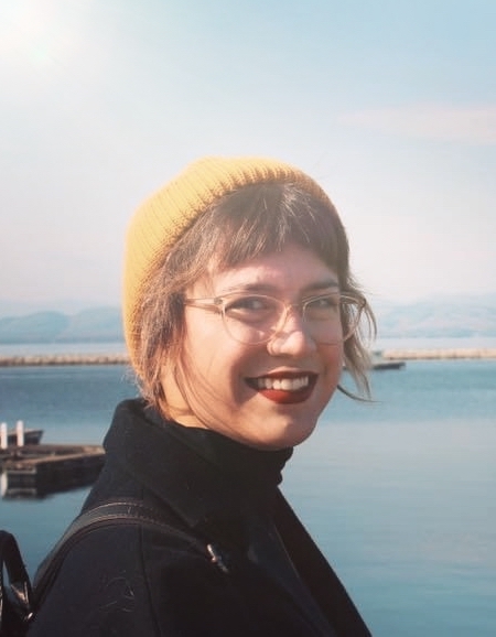 Image description: Kendra is a White, femme-presenting person with brown hair and bangs, wearing a yellow tuque, a blue jacket, clear-framed glasses, and red lipstick. She is looking over her right shoulder smiling. Behind her is a blue sky and blue water, a pier, and mountains in the distance at downtown Burlington, Vermont.