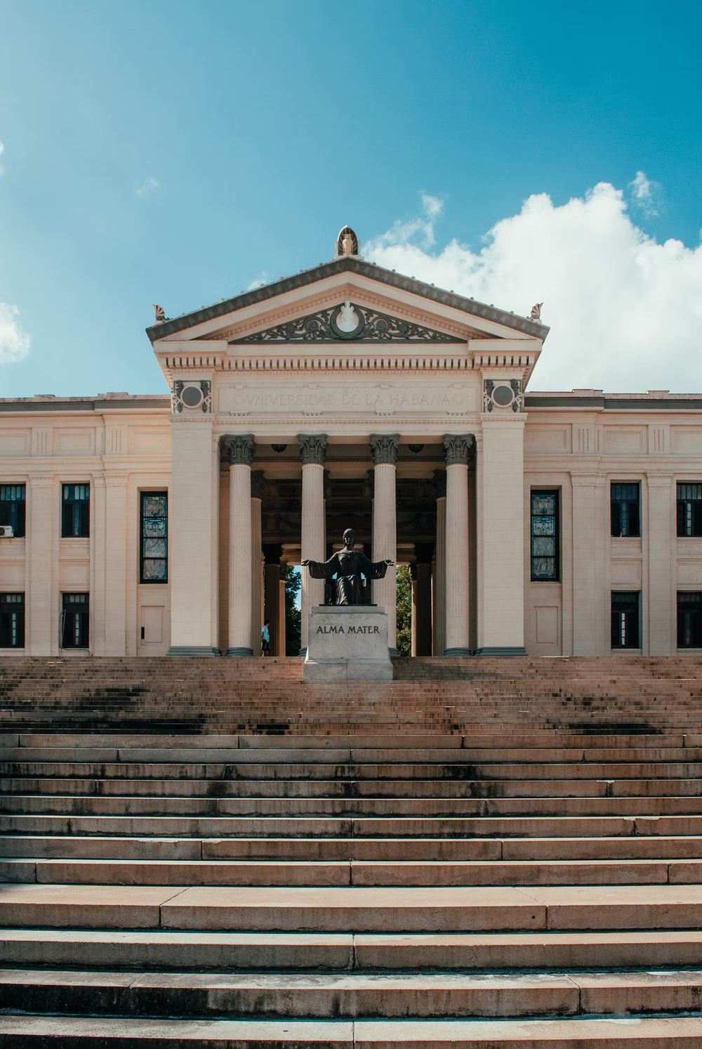 Image description: A photo of steep steps at the University of Havana, Cuba. At the top of the steps is a replica of the Alma Mater, also known as the "nourishing mother." The figure, clad in a bulky gown and crowned with a laurel wreath, sits on a throne with both arms mid-raised. A large book, representing knowledge, sits open on her lap. The original sculpture of the Alma Mater is located at the top of the steps to Low Memorial Library on Columbia University campus in New York City. Photo by Remy Gieling.